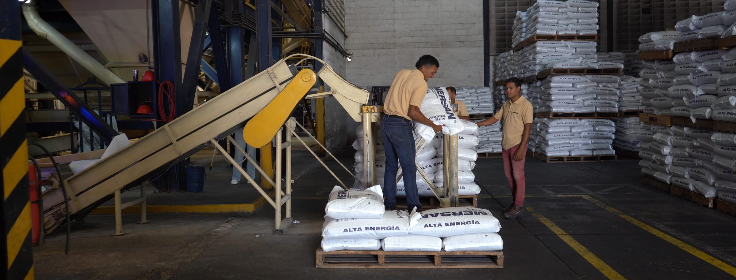 Dos operarios paletizando sacos de alimento Mersan junto a la cinta de ensacado, con palés apilados al fondo